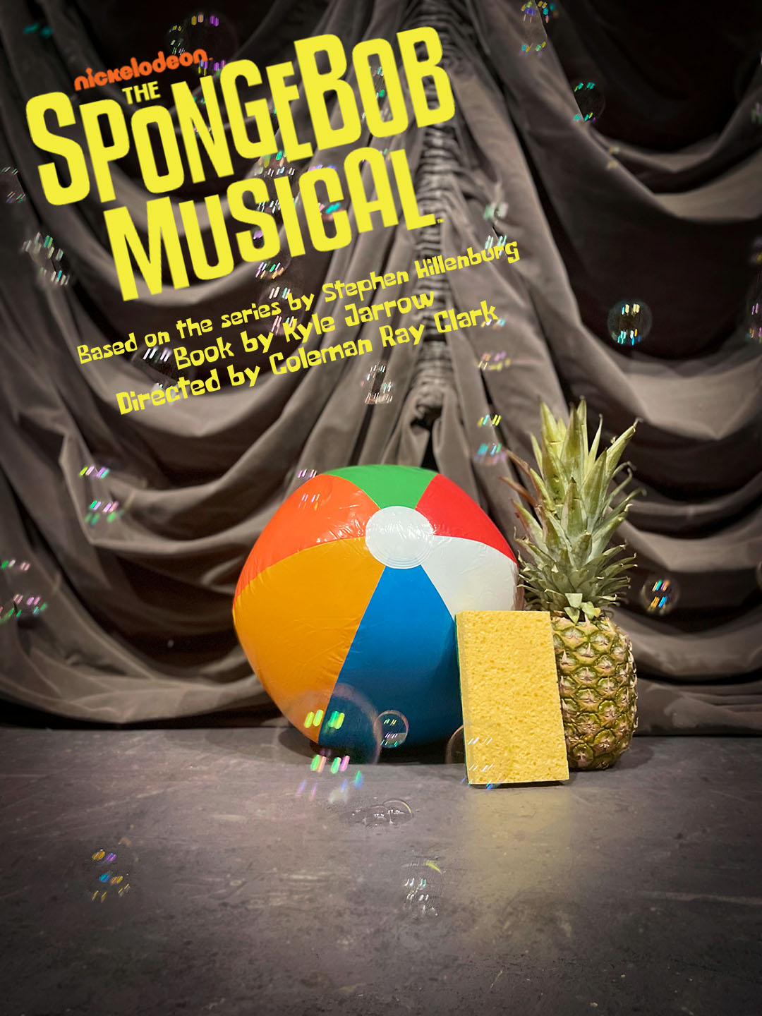 A pineapple, a beach ball, and a yellow sponge are arranged in front of a theatrical drape on a s...